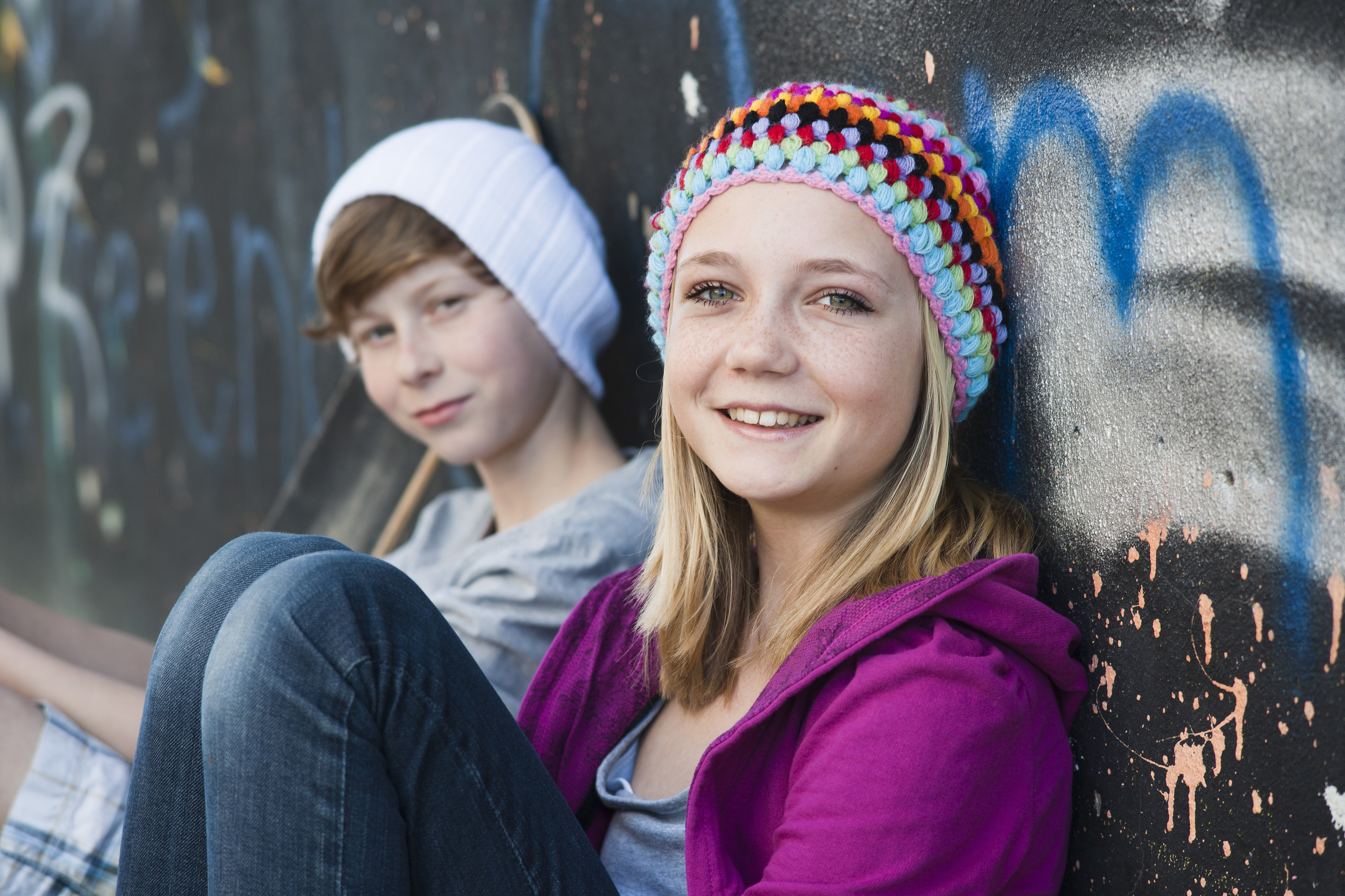 Illustrassjonsbilde, Jente og gutt, ca 12 år, sitter inntil vegg med graffiti, smiler og ser i kamera.