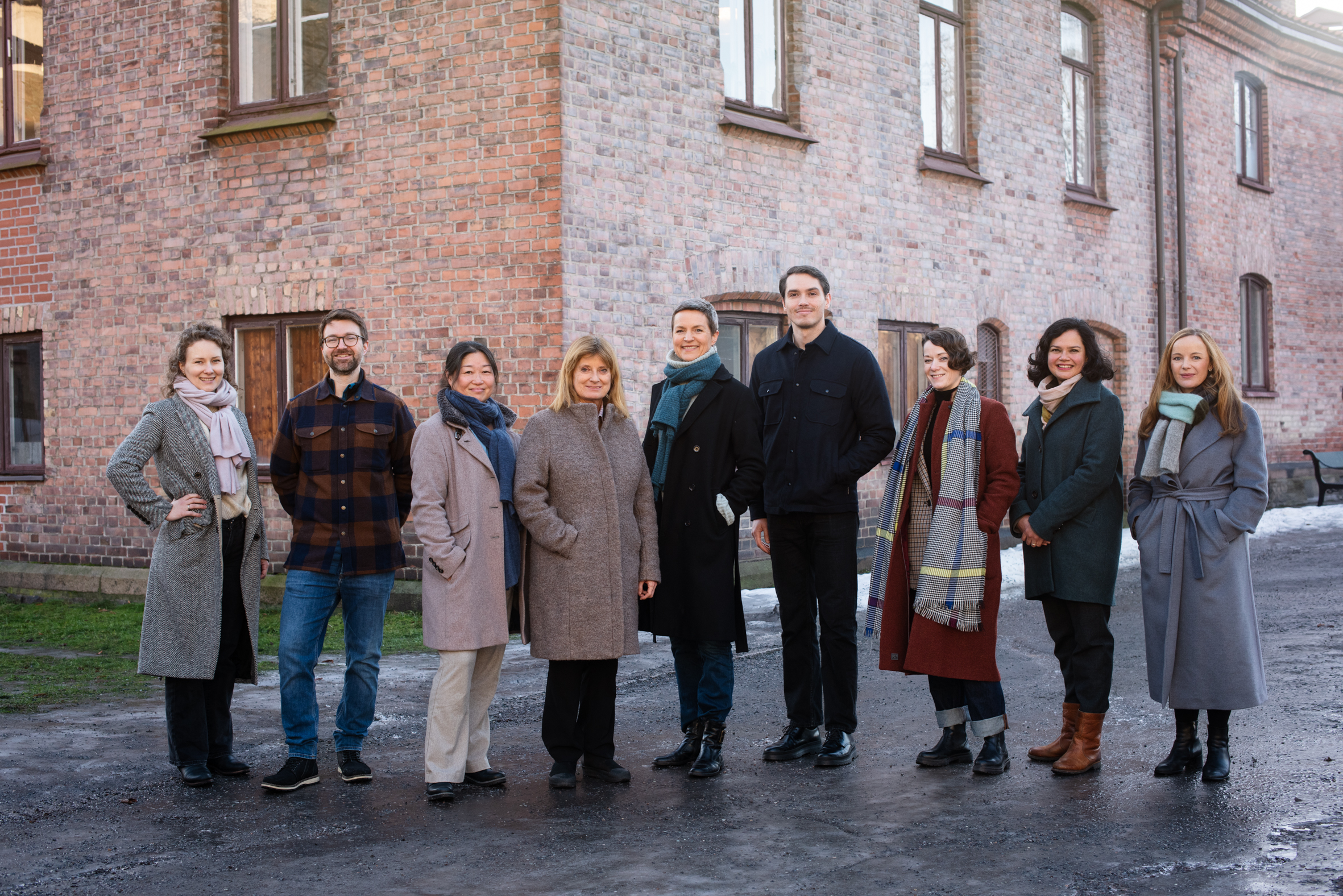 Gruppebilde av ni personer i yttertøy, fotografert utendørs foran lys murbygning