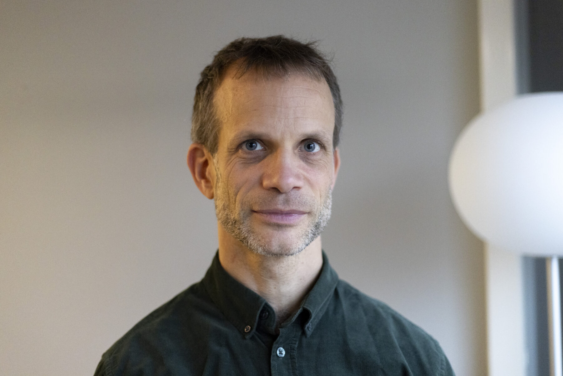 Portrait photo of a man wearing a dark shirt, white round lamp to the right in the background.