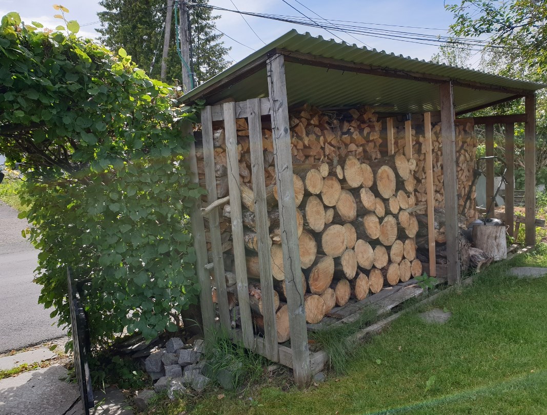 Vedskur med ved stablet opp. Grønt gress foran, grønn busk til venstre.