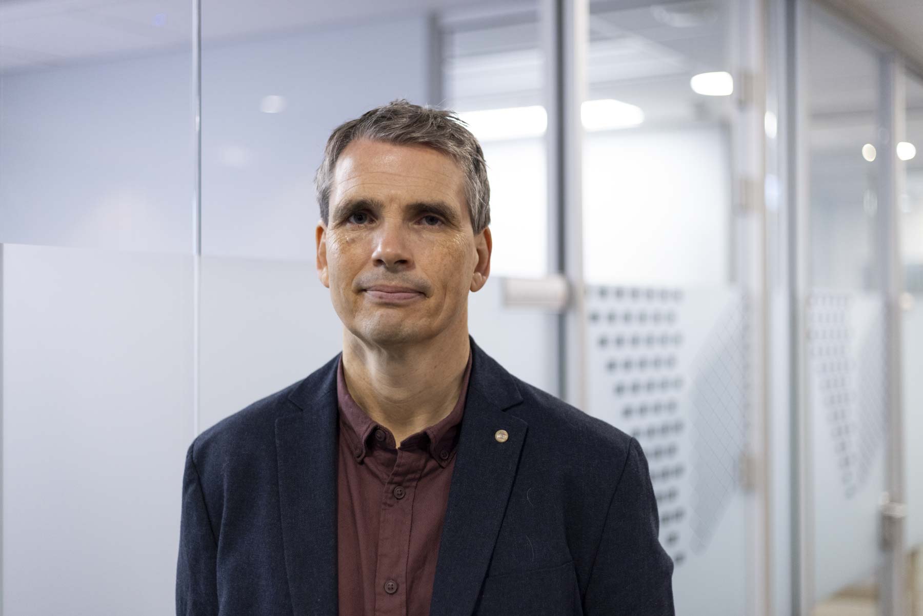 Portrettfoto mann, burgunder skjorte og mørk blå jakke, mot bakgrunn av glassvegger.