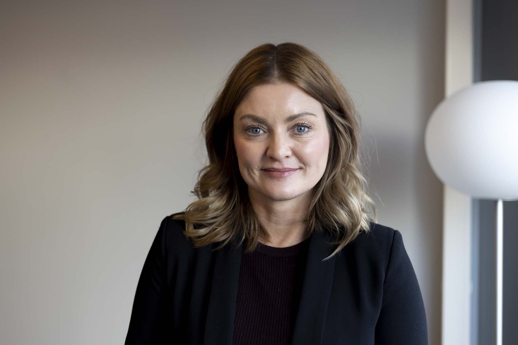 Portrettfoto kvinne i mørkt antrekk, halvlangt hår, hvit rund lampe til høyre i bakgrunn.