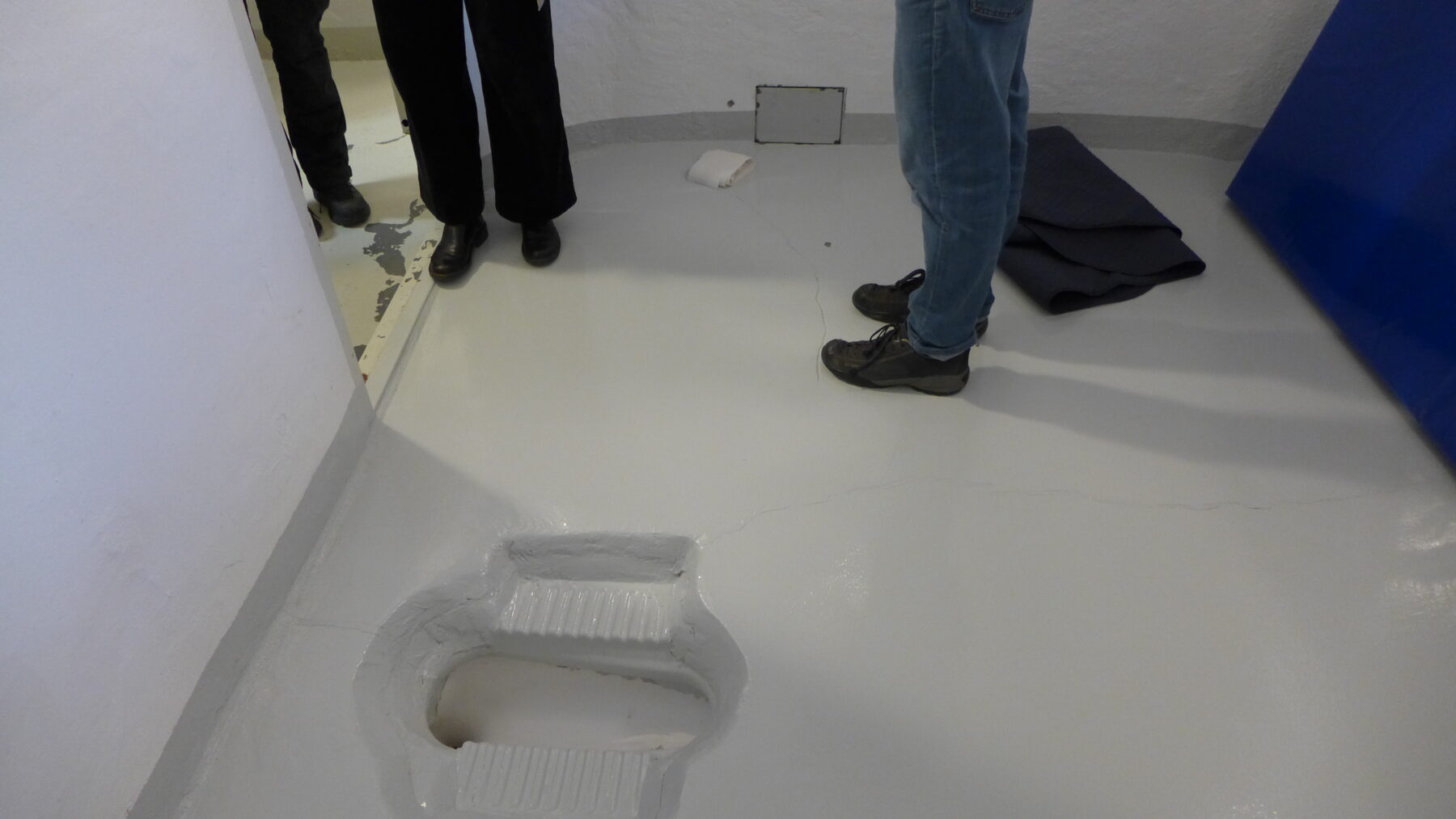 Golvet i en sikkerhetscelle, fotografert med tre par føtter i bildet