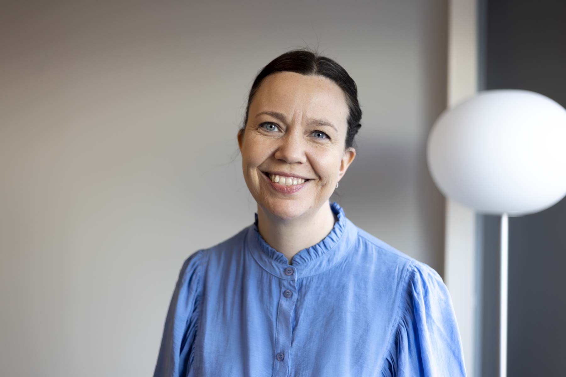 Portrait photo of a woman with dark hair, wearing a light blue blouse, round white lamp in the background.