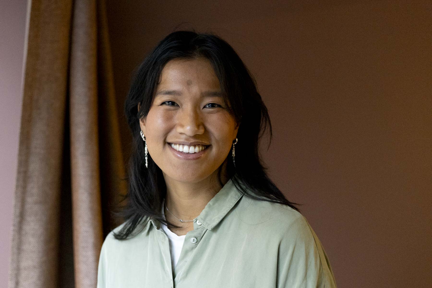 Portrettfoto dame med lys turkis skjorte. mørkt halvlangt hår, brunt gardin og vegg i bakgrunn